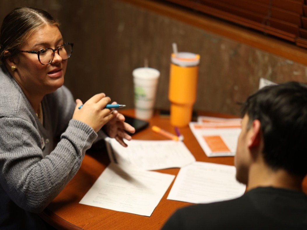 A brunette with a ponytail is wearing glasses and talking to a student, who has short dark hair. 