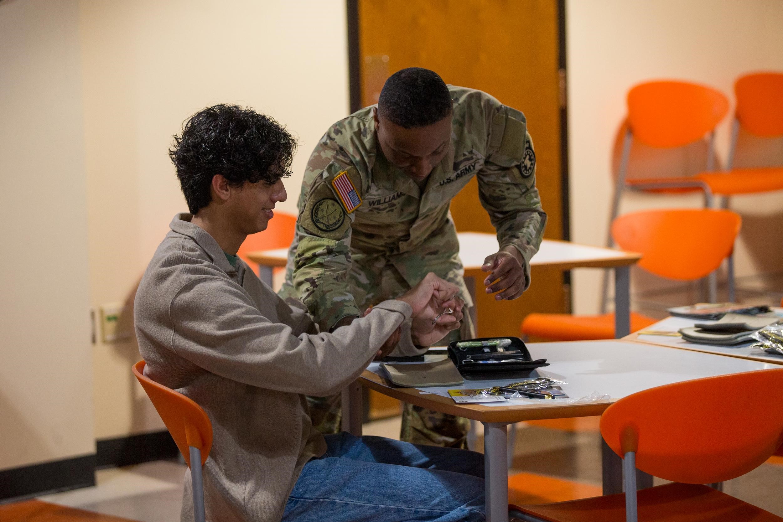 A man in a camouflage uniform is leaning over a student working with his hands at a suture clinic.