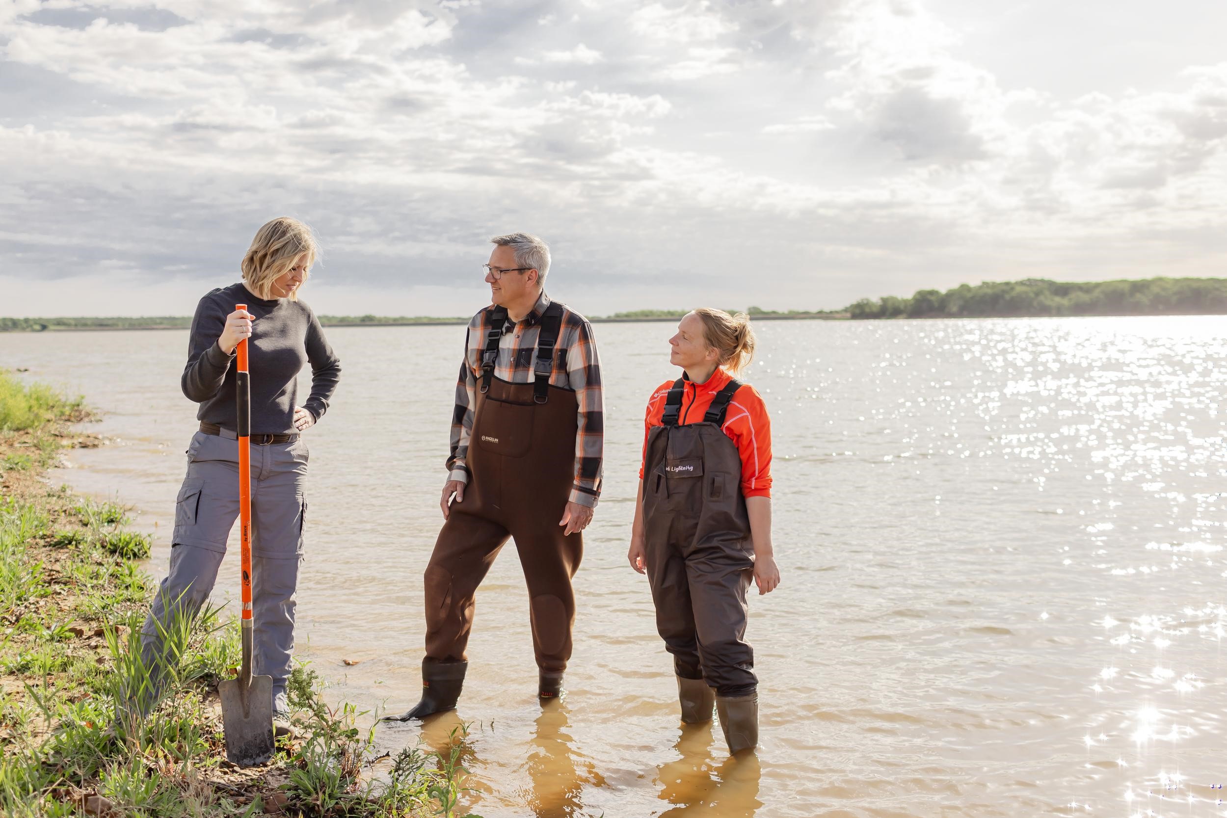Two women and a man are standing in the shallow part of a lake researching the water quality.