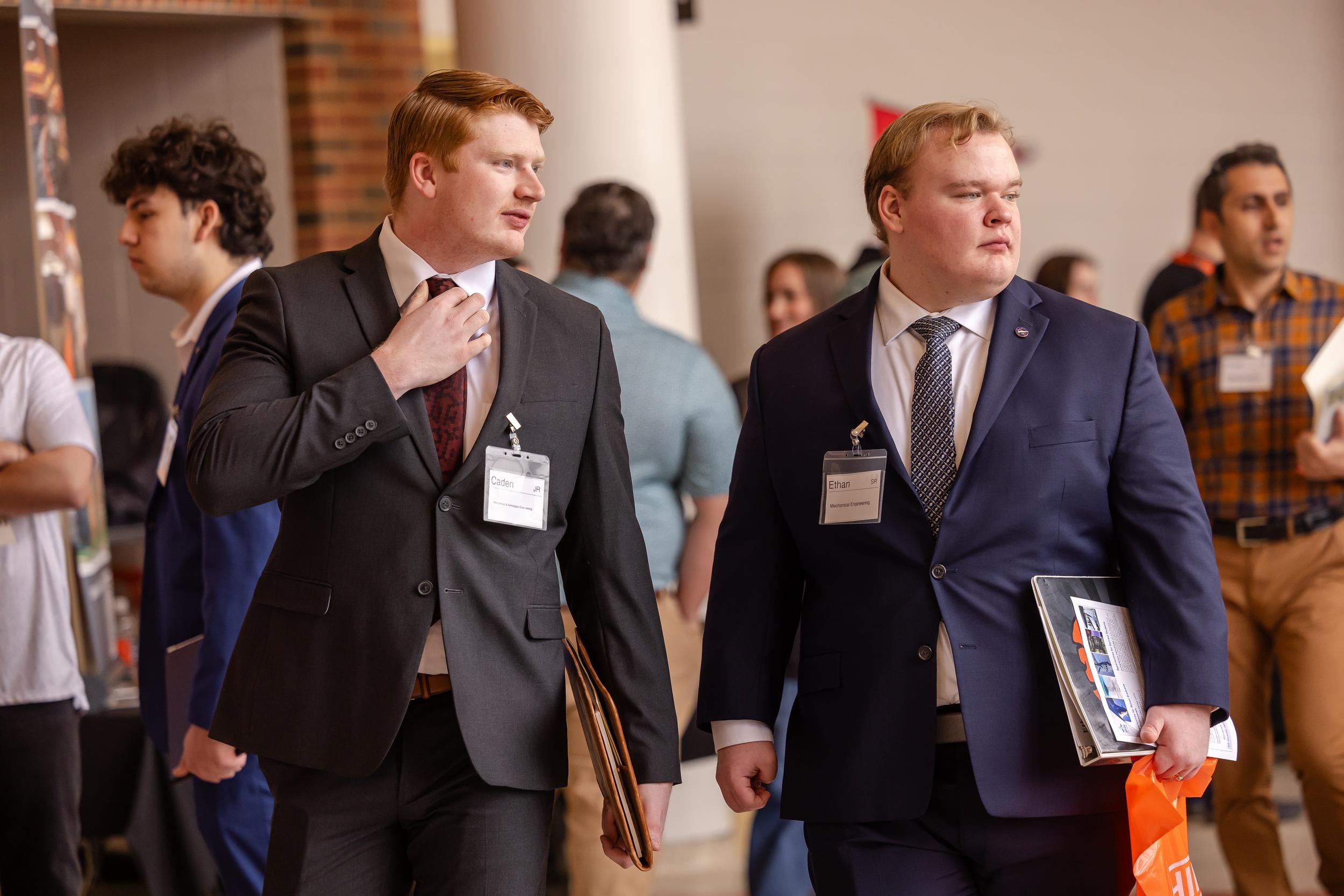Two students walk through the career fair. One man is adjusting his tie. They are both dressed in suits.