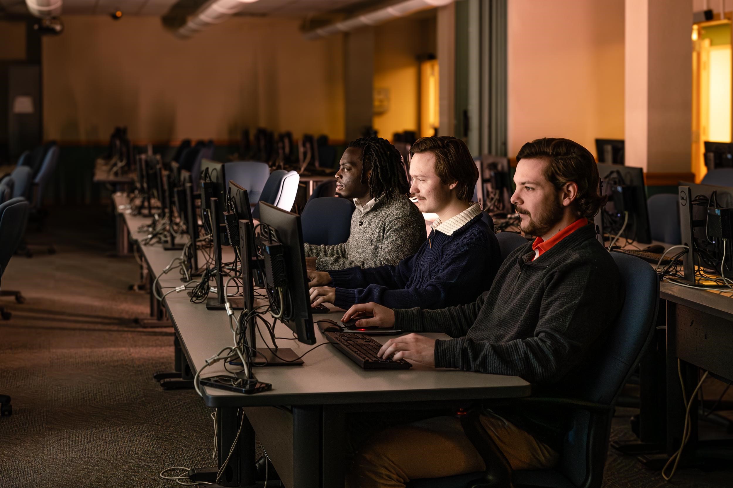 Three men sit smiling and facing computers in a dark room lit only by the computer screens