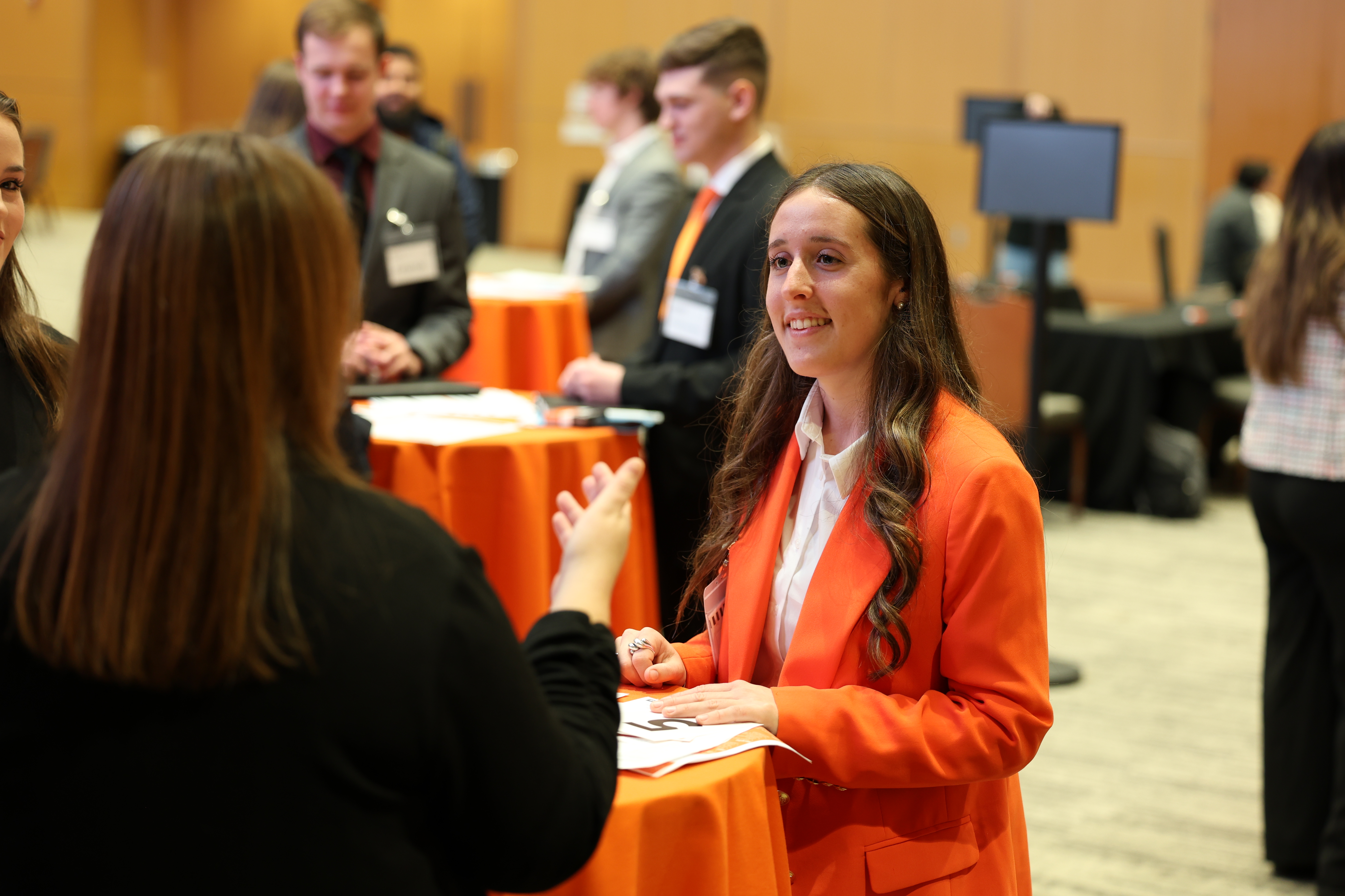A brunette student in an orange blazer is talking to a staff member who is turned away from the camera. They are at a Career Services networking event. 